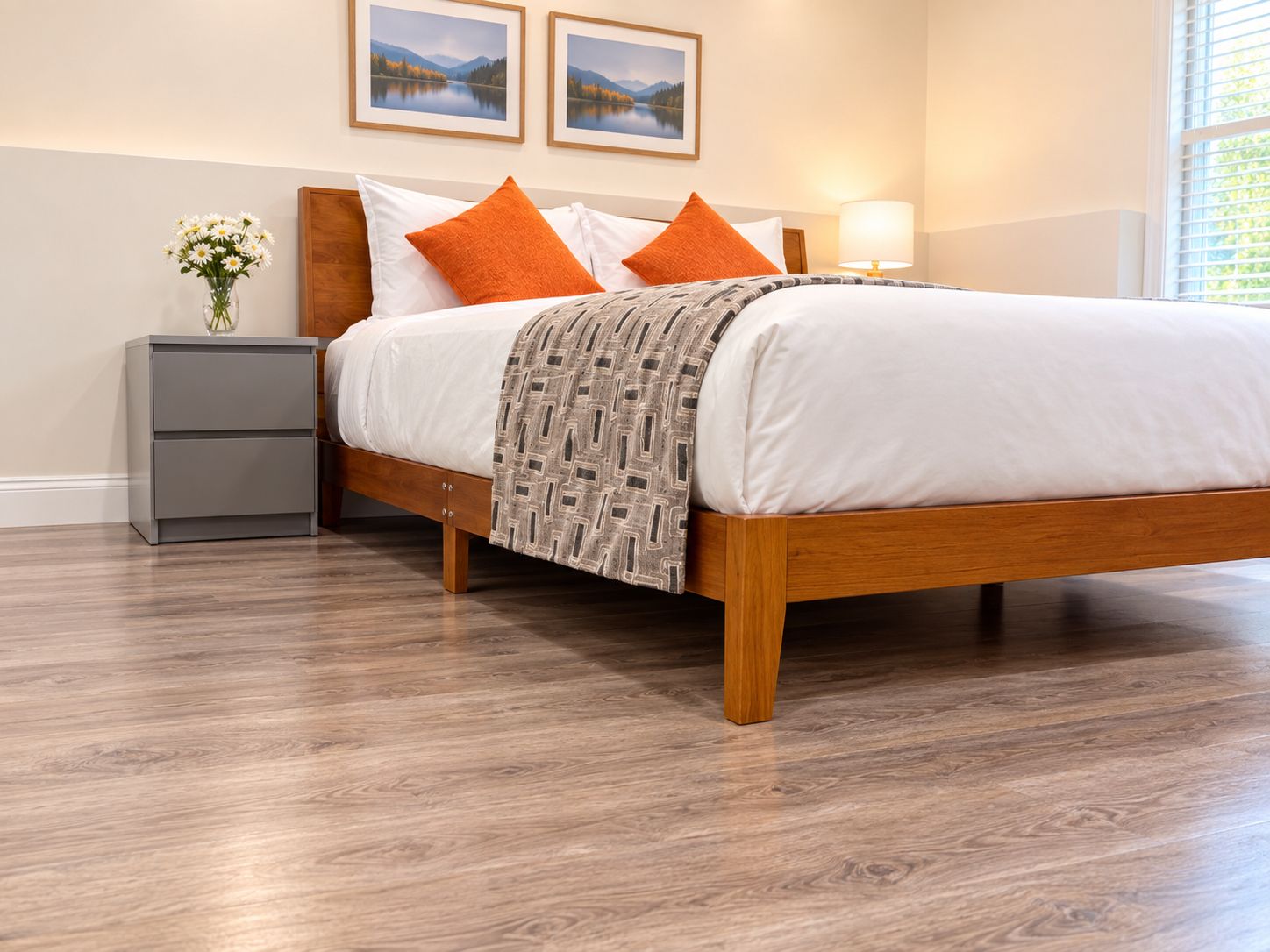 A bedroom with a wooden bed, white bedding, and two orange pillows. There is a bedside table with a lamp, a gray nightstand, and a vase with flowers. Two framed pictures are on the wall above the bed.