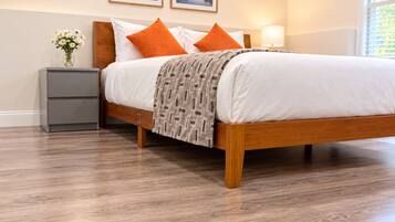 A bedroom with a wooden bed, white bedding, and two orange pillows. There is a bedside table with a lamp, a gray nightstand, and a vase with flowers. Two framed pictures are on the wall above the bed.