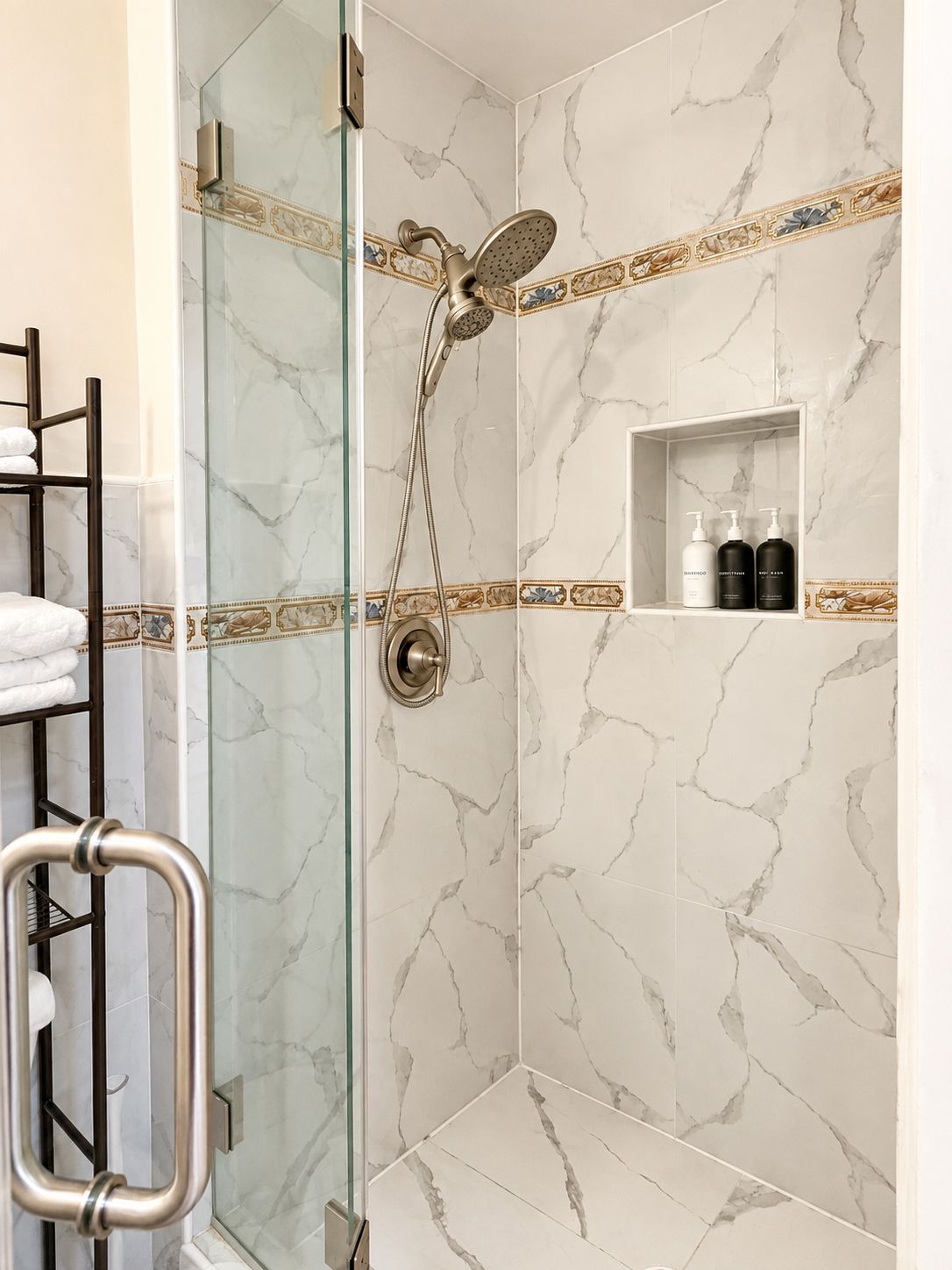 A shower with a glass door, marble walls, and a shelf with toiletries.