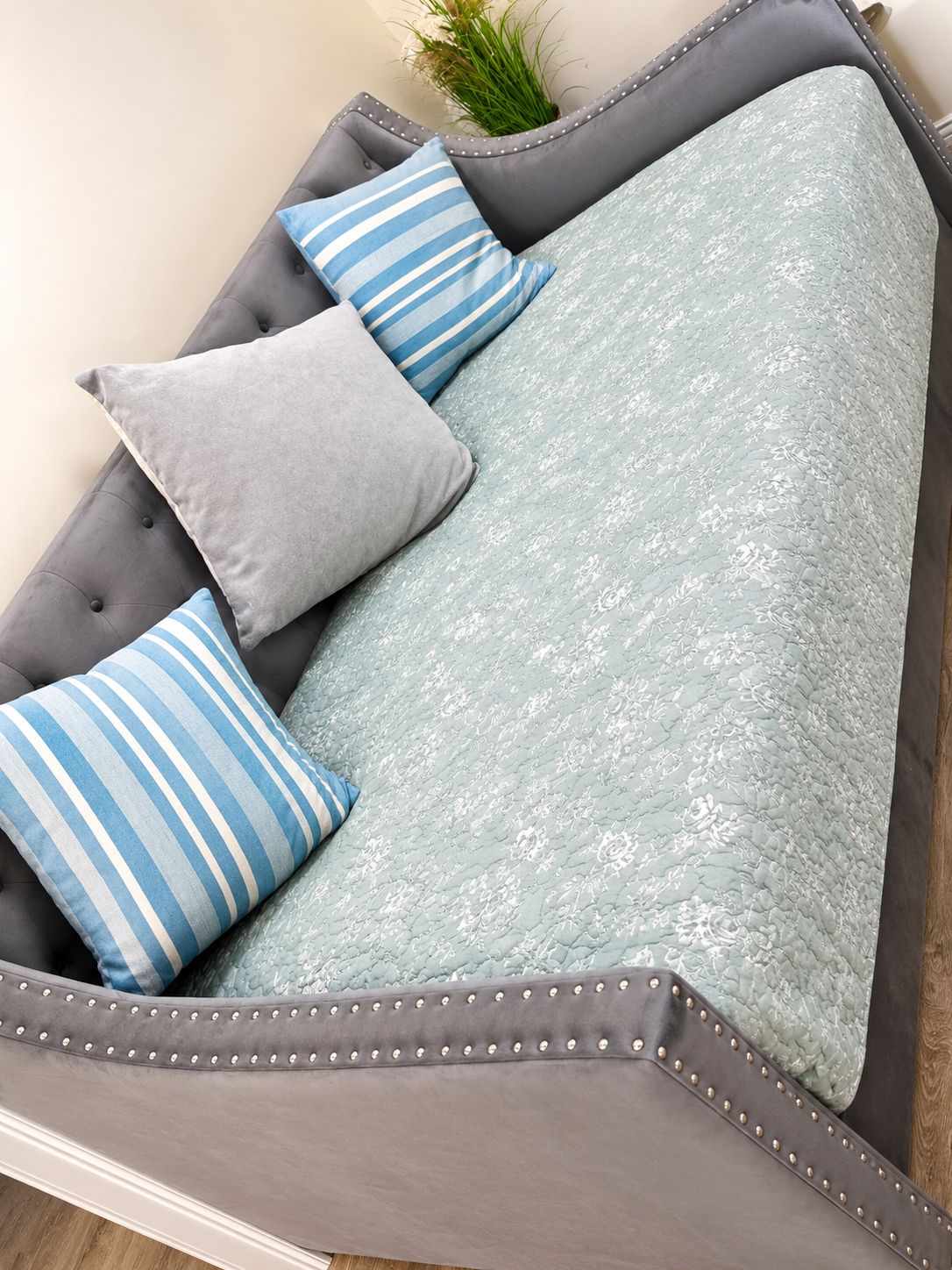 Ein graues, gepolstertes Sofa mit einem floralen Bezug und vier Kissen, davon ein gestreiftes und drei einfarbig grau.