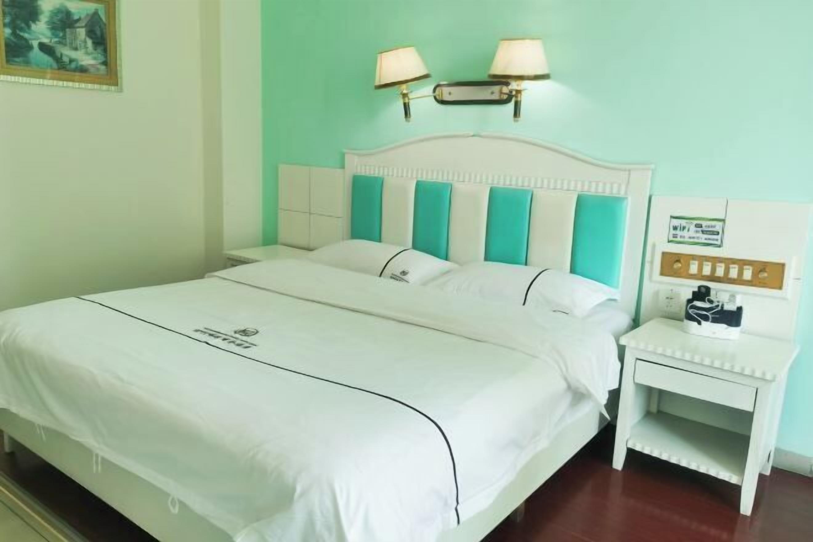 Un lit bien fait, avec une literie blanche et des coussins turquoise, une table de chevet avec une lampe et un luminaire fixé au mur.