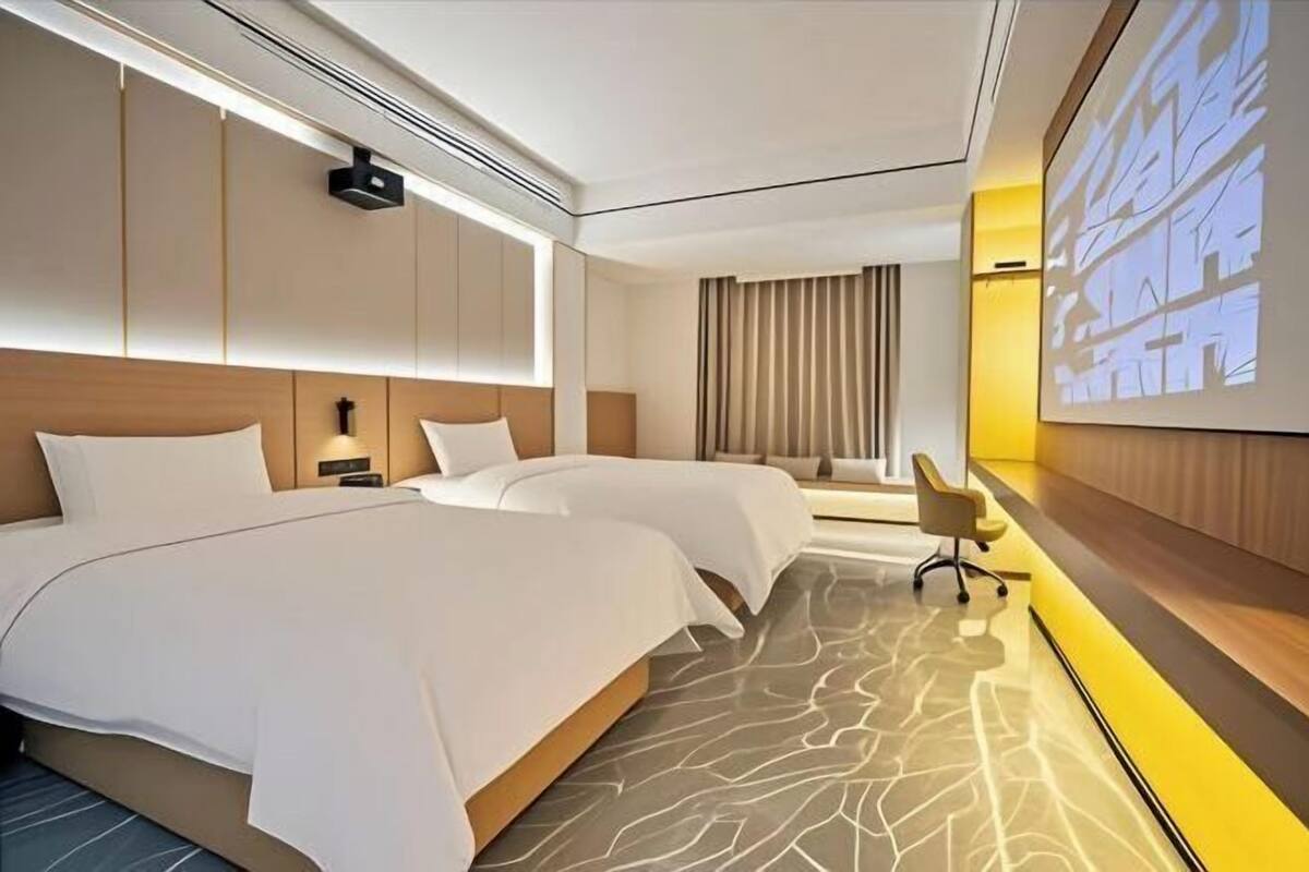 Ein modernes Hotelzimmer mit zwei Betten, einem Schreibtisch, einem Sessel und einem großen Bildschirm, der eine Stadtansicht zeigt.