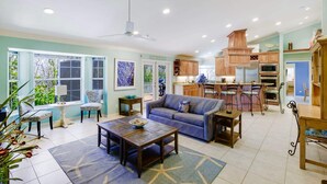 A spacious living area with a sofa, coffee table, and chairs, a kitchen with wooden cabinets and appliances, and large windows with a view of greenery. - Gorgeous 5 Bedroom Home with Heated Pool and Spa (Sanibel)