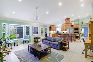 A spacious living area with a sofa, coffee table, and chairs, a kitchen with wooden cabinets and appliances, and large windows with a view of greenery.