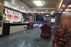 Zona de recepción de un hotel, con mostrador de recepción, sillas y una alfombra roja que conduce a la entrada.