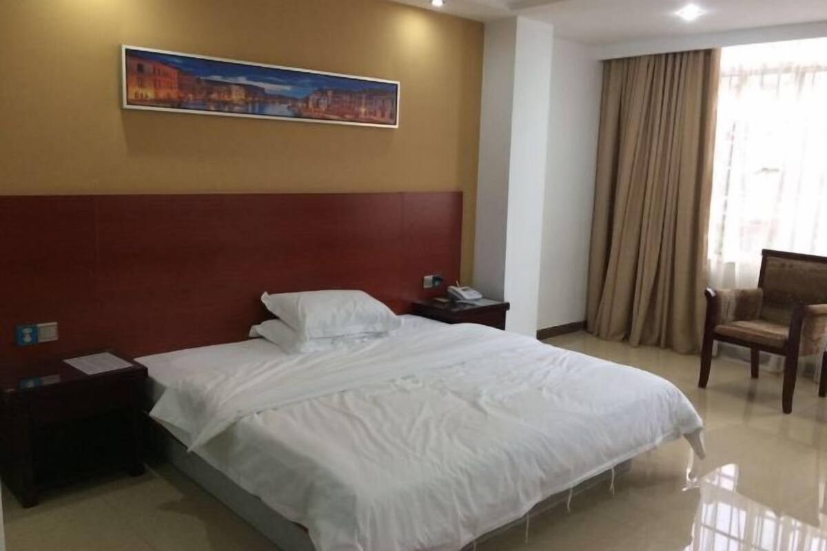 A hotel room with a large bed, a nightstand, a chair, a painting on the wall, and a window with curtains.
