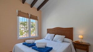 A bedroom with a large bed, two windows with blinds, a bedside table with a lamp, and a view of buildings outside.