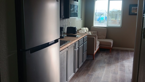Een compacte keuken met een koelkast, magnetron en een klein aanrecht. Er is een raam met zonwering, een bank en een deur naar een andere kamer.
