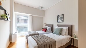 Quarto moderno com duas camas, uma mesinha e uma janela com persianas.