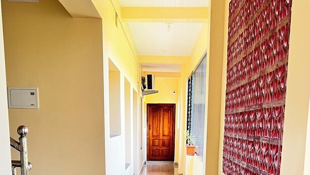 A hallway with a tiled floor, a wooden door at the end, and a red decorative wall on the right. - MM Garden (Tarangambadi)