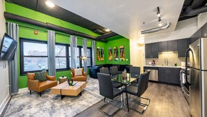 Un appartement moderne avec un mur d’accent vert, un coin salon confortable avec un canapé et des fauteuils, une table à manger et une cuisine équipée d’appareils électroménagers en acier inoxydable.