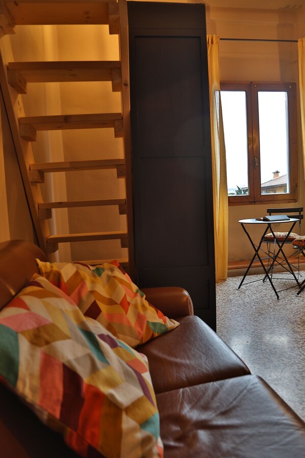 A room with a brown leather sofa, a wooden ladder, a small table with a chair, and a window with curtains.