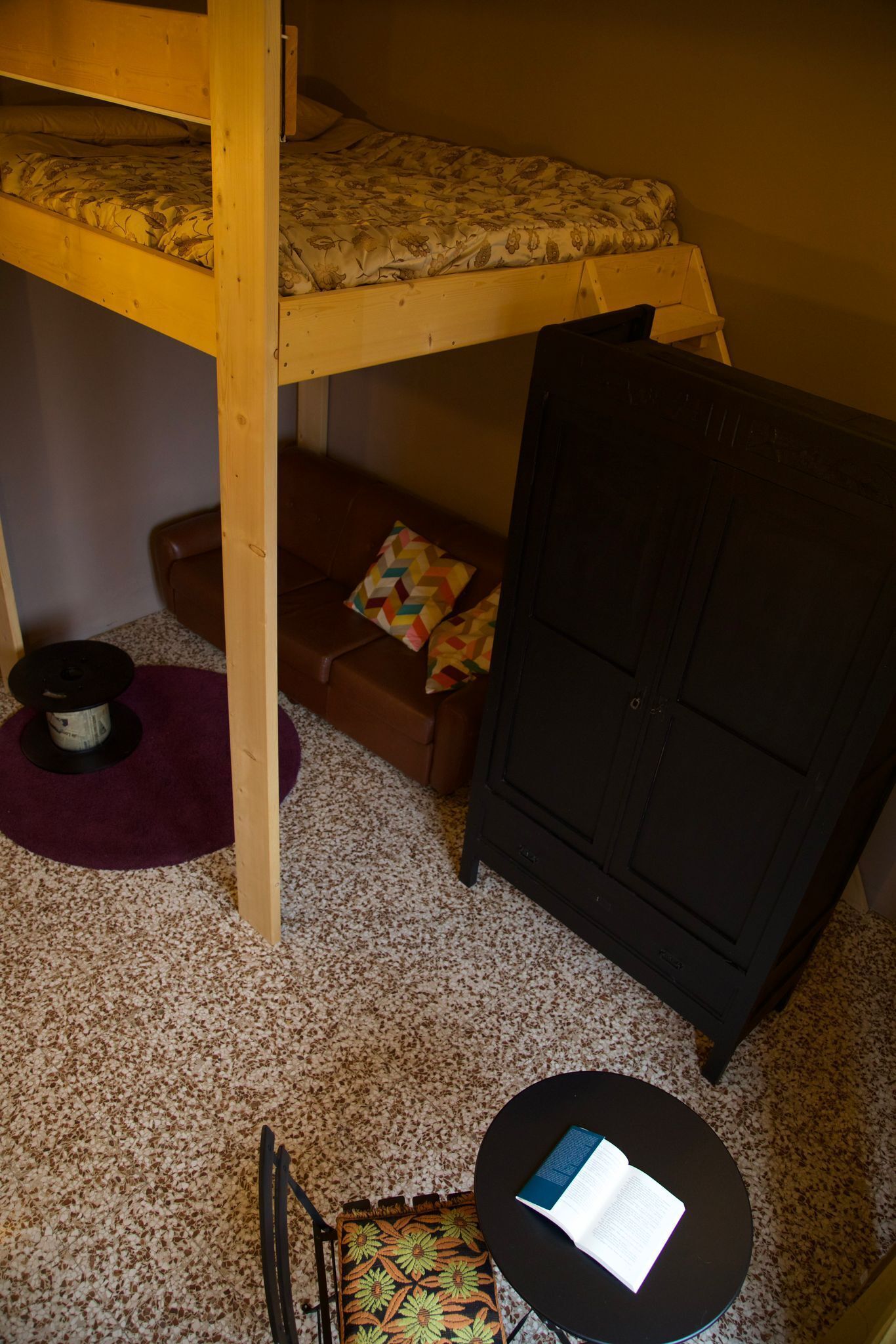 A bunk bed with a patterned bedspread, a brown leather sofa with a colorful pillow, a black cabinet, a round table with a book, and a spool of thread on a purple rug.