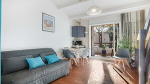 A living room with a grey sofa, a dining table with chairs, a TV, and a large window with a view of trees. - Bright apt for 4 ppl, balcony, private parking (Agde)