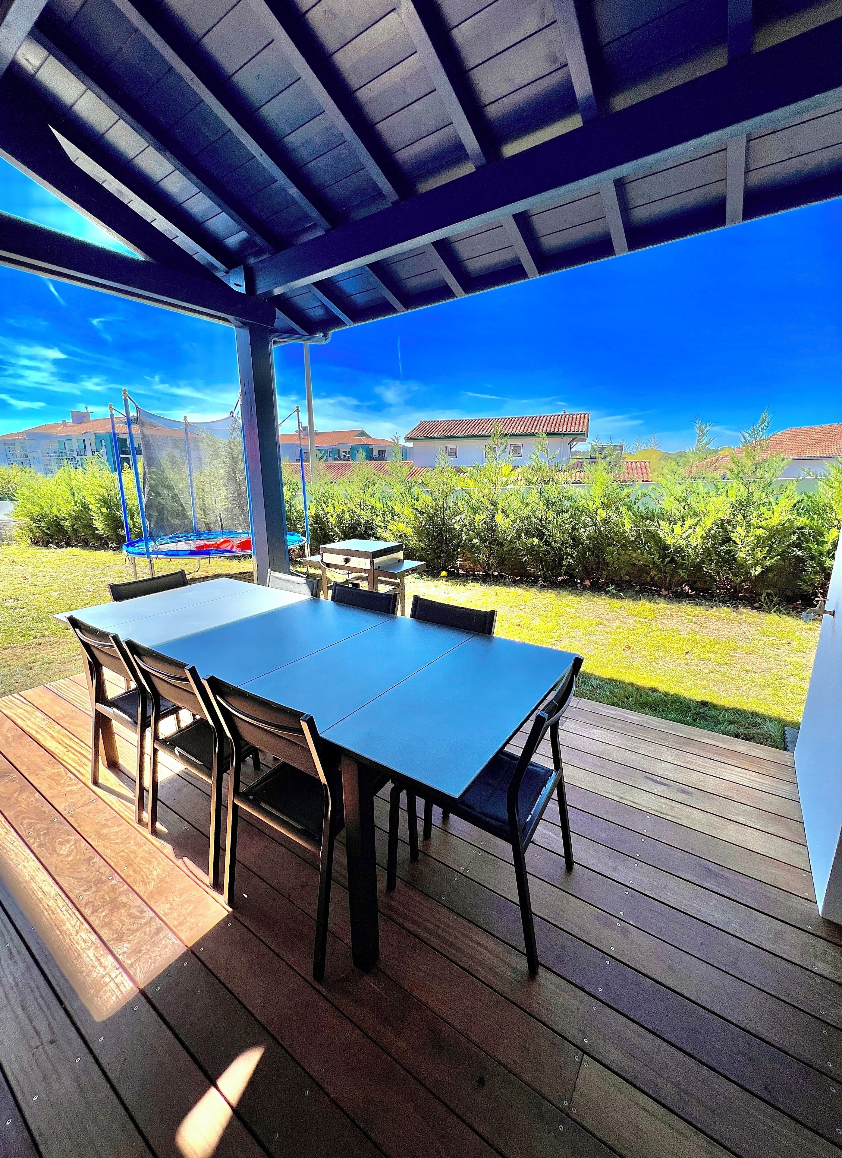 Un patio exterior con mesa y sillas, un trampolín y vistas a casas y vegetación.