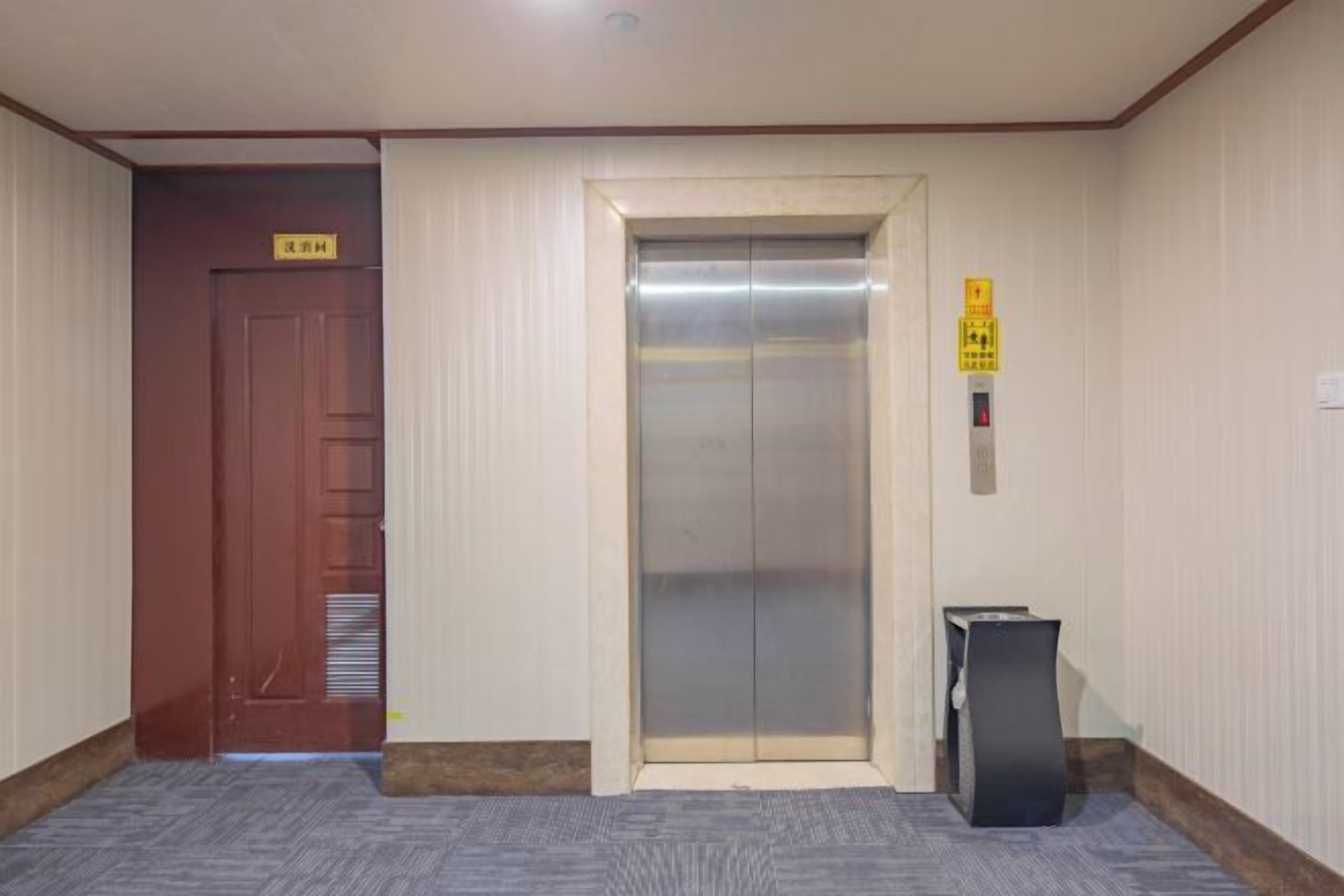 A hotel elevator with a sign above it, a wooden door to the left, and a trash bin to the right.
