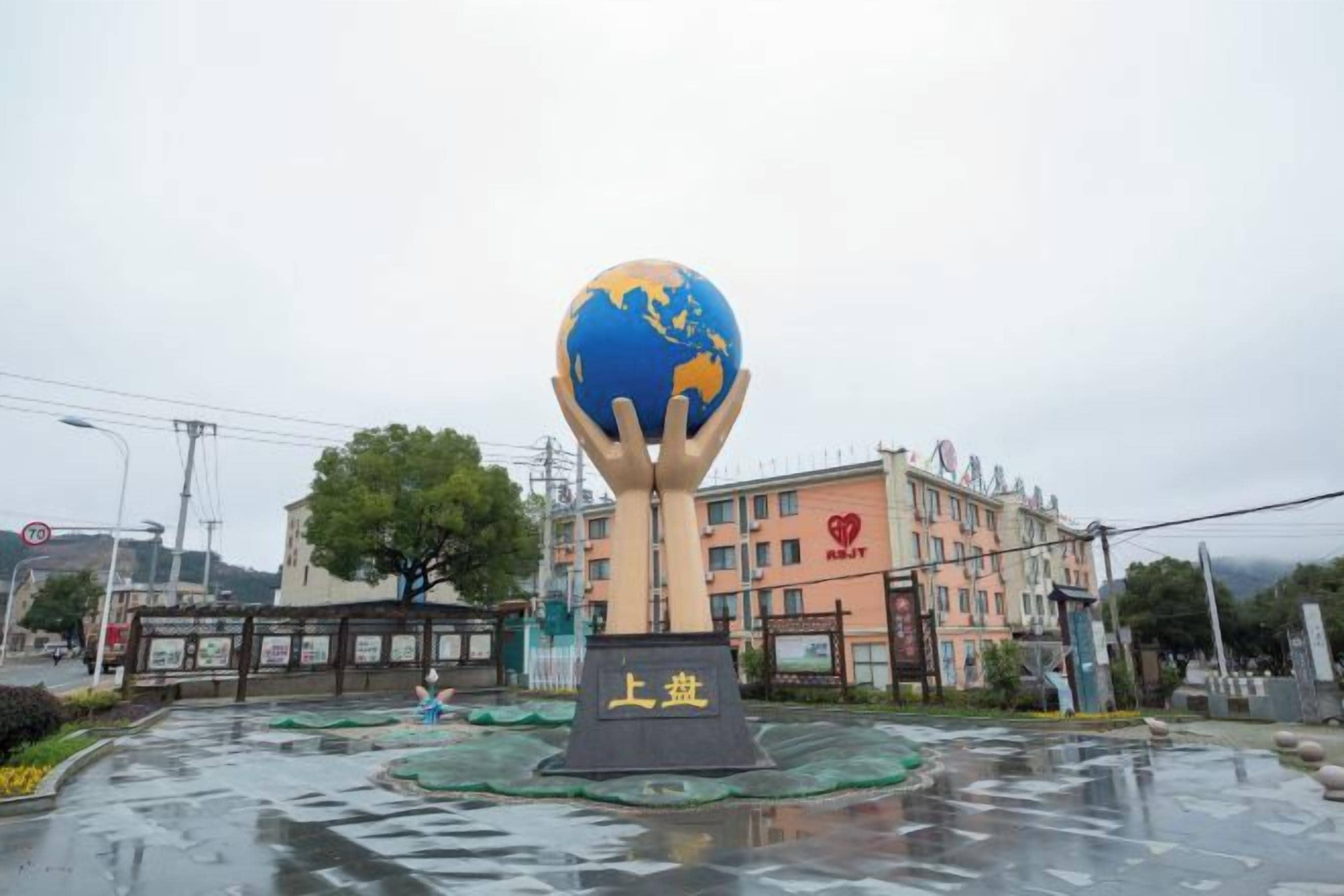 Une sculpture représentant un globe terrestre, surmontée d’une plaque, entourée d’un pavé mouillé, avec des bâtiments en arrière-plan.