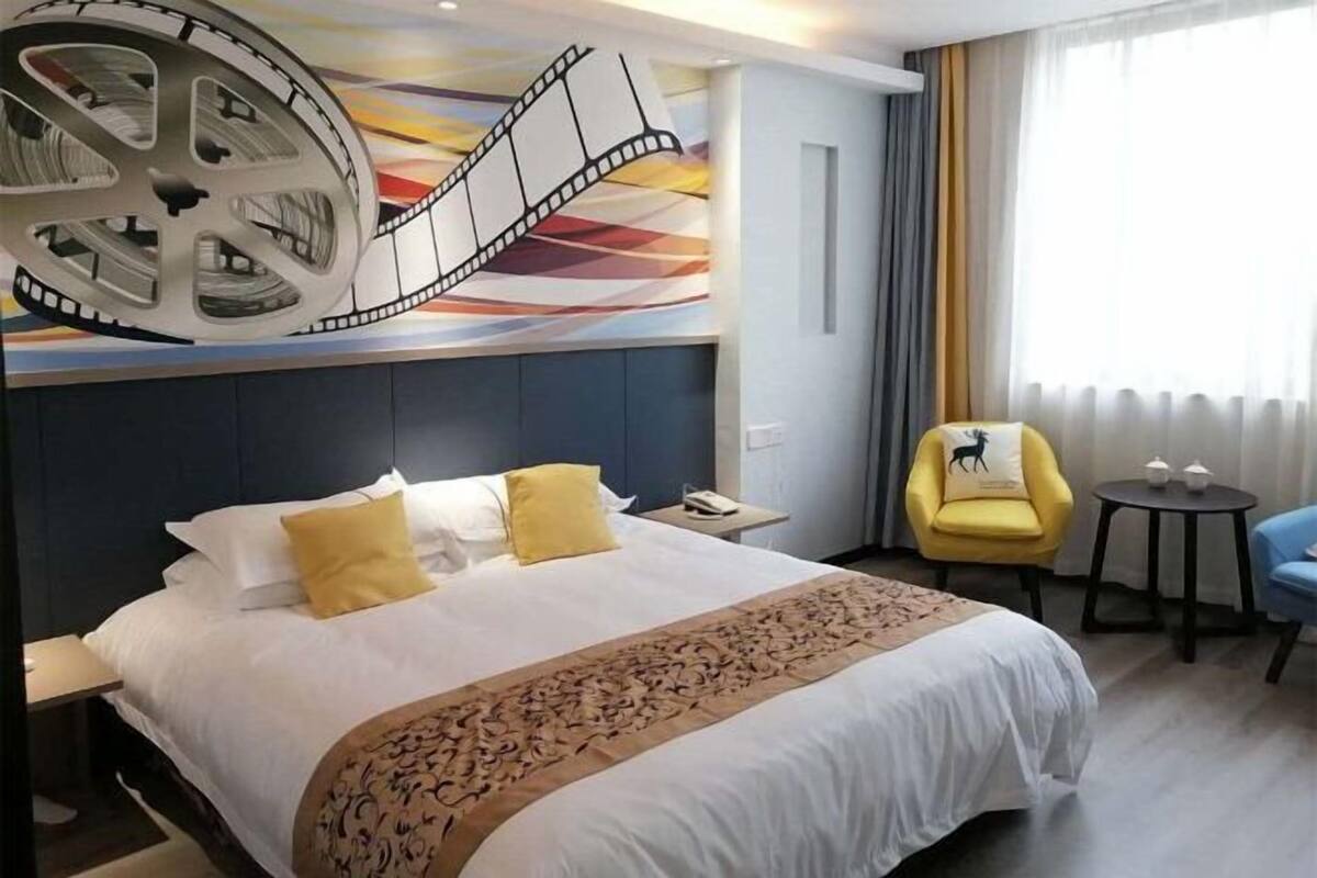 A hotel room with a large bed, a headboard with a film reel design, a small table, a chair, and a window with curtains.