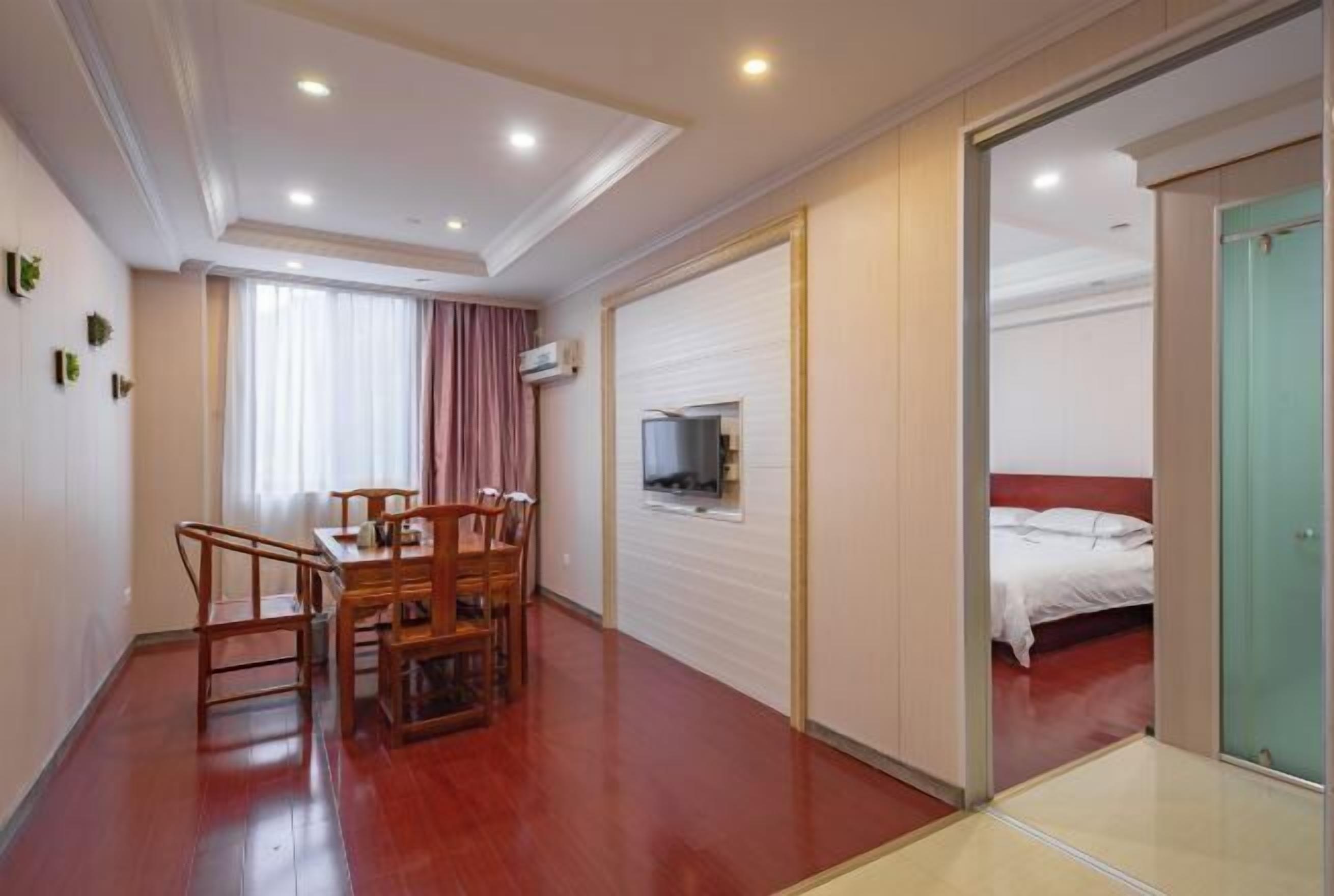 A hotel room with a wooden dining table, chairs, a bed with white bedding, a television mounted on the wall, and a sliding door leading to another room.