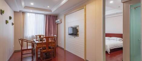 A hotel room with a wooden dining table, chairs, a bed with white bedding, a television mounted on the wall, and a sliding door leading to another room.