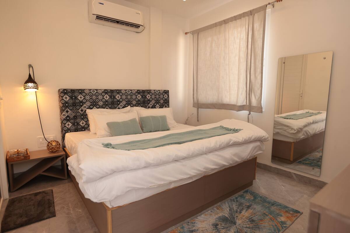 A bedroom with a bed, bedside table, lamp, air conditioning unit, window with curtains, and a mirror.