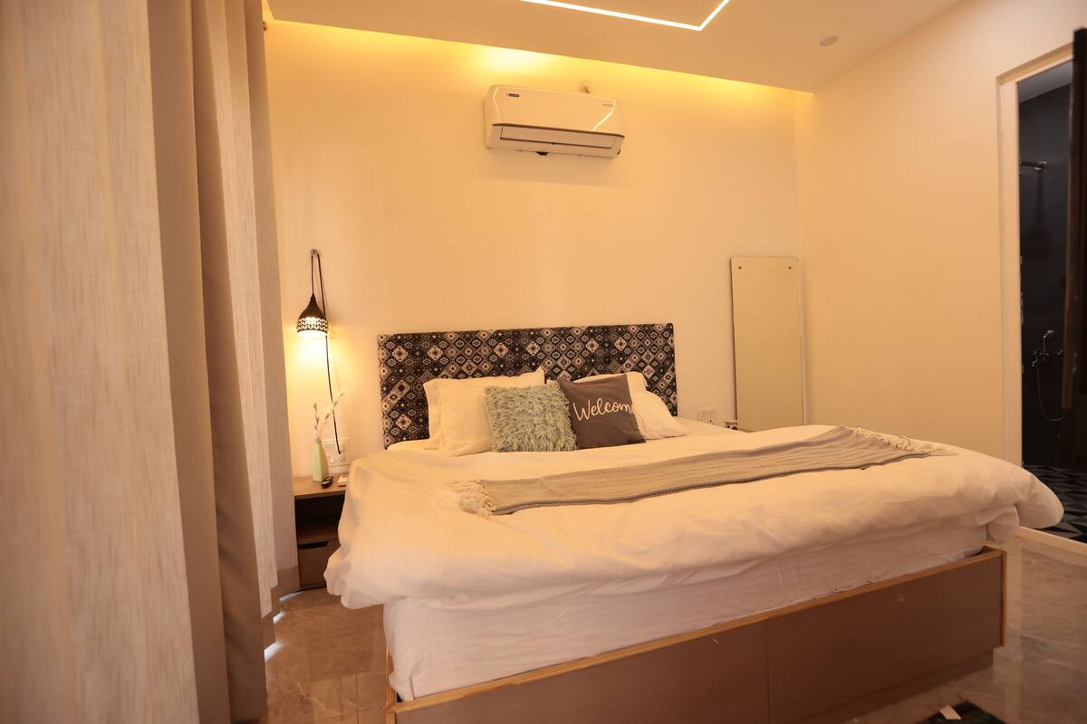 A neatly made bed with a patterned headboard, a bedside lamp, and a wall-mounted air conditioner.