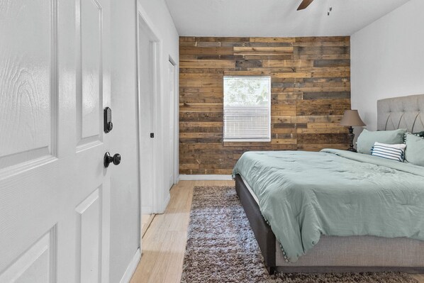 Une chambre avec un mur d’accent en bois, un lit recouvert d’une courtepointe verte, une table de chevet avec une lampe et une fenêtre avec des stores.