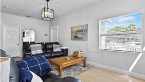 A living room with a black leather sofa, a wooden coffee table, and a large window with blinds. - 4BR Charming Home By Beachside (West Palm Beach)