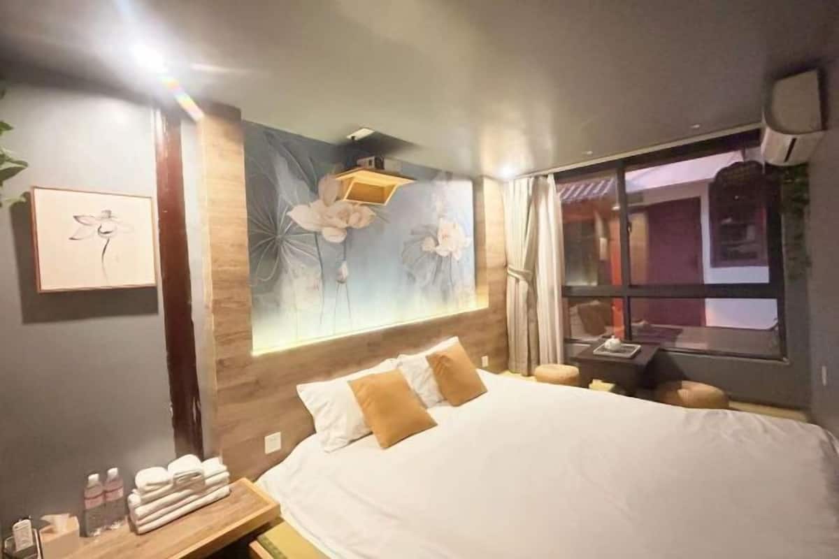 Ein Hotelzimmer mit einem großen Bett, einem Holzkopfende, einem Wandbild, einem Nachttisch mit Toilettenartikeln und einem Fenster mit Vorhängen.