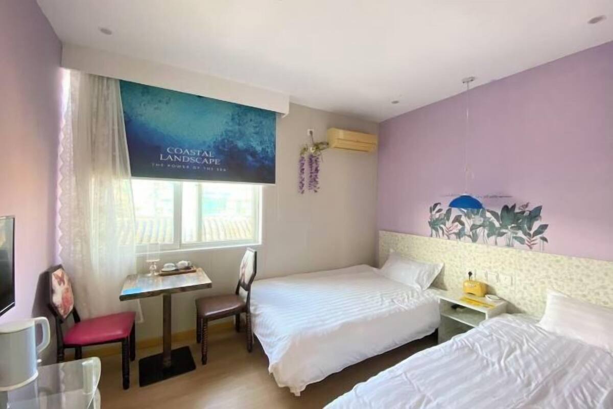 Ein Hotelzimmer mit zwei Betten, einem kleinen Tisch, einem Stuhl, einem Fernseher und einem Fenster mit Vorhängen.