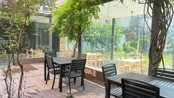 An outdoor patio with tables and chairs, surrounded by glass walls and greenery.