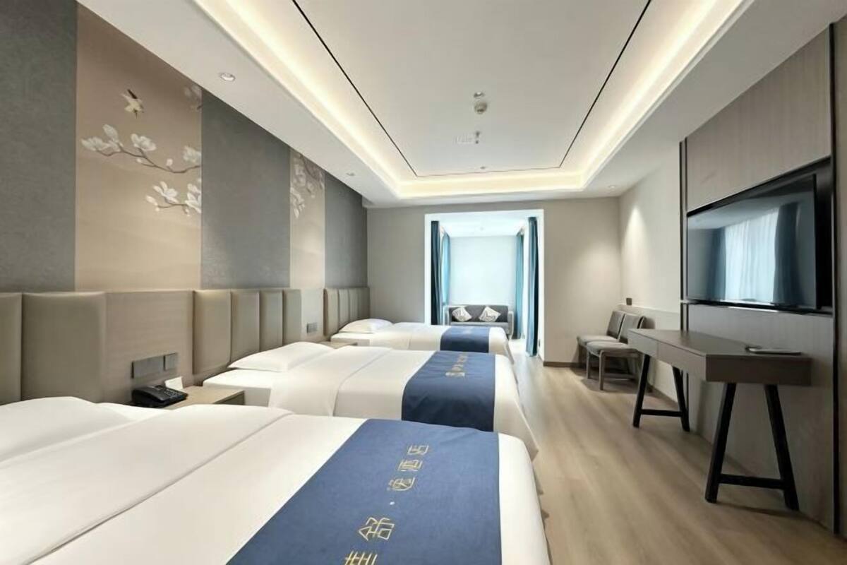Habitación de hotel con tres camas, un escritorio, un televisor y un ventanal grande.