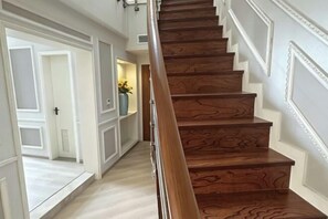 Una escalera de madera con barandilla, paredes blancas con molduras decorativas y un pasillo que conduce a una puerta con un símbolo de cruz.
