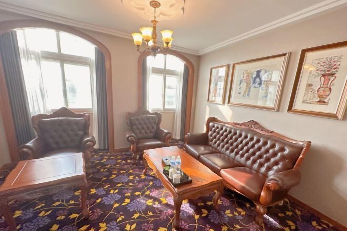 Una habitación con alfombra floral, una mesa de centro de madera, un sofá de cuero, dos sillones, una araña y cuadros en la pared.