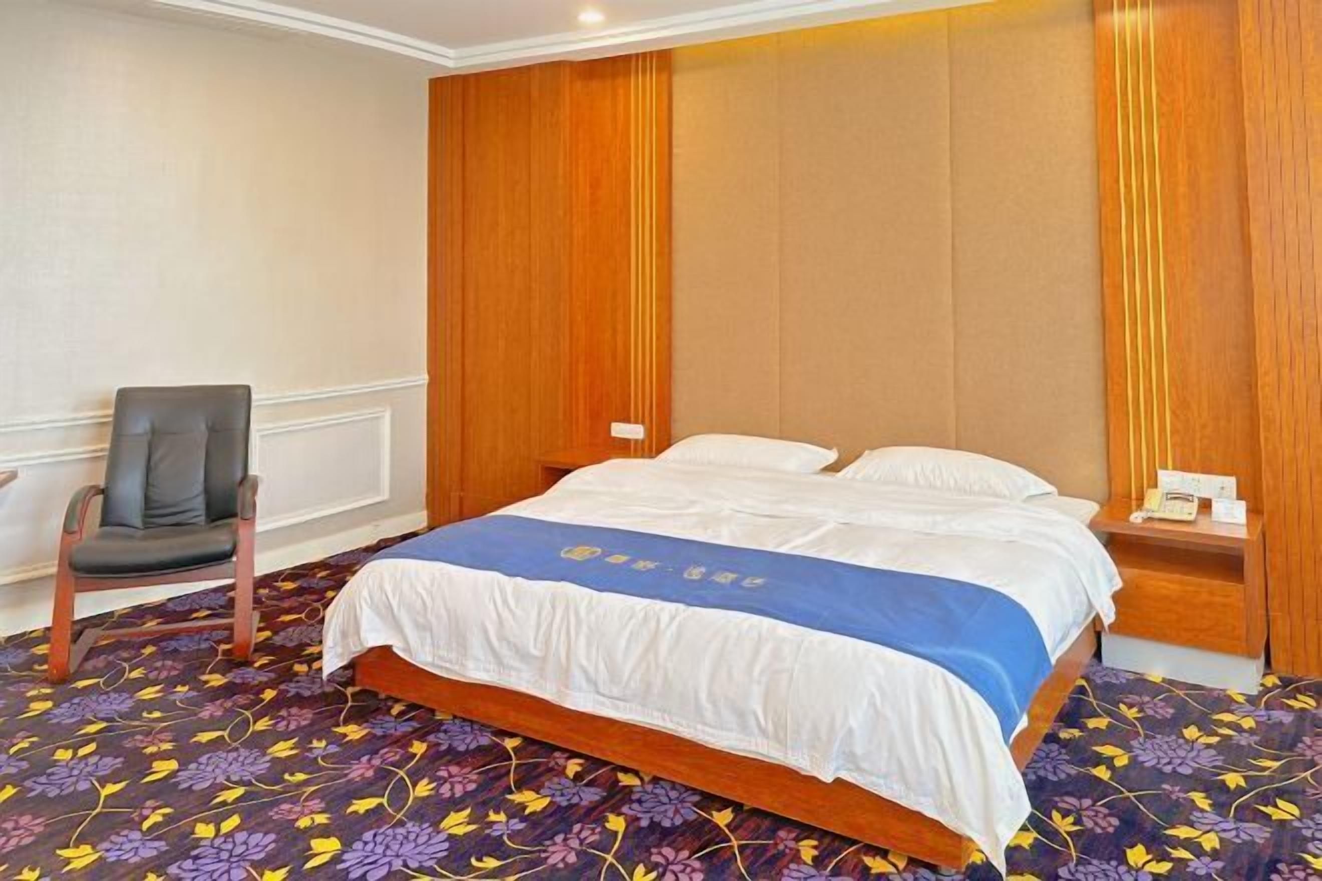 A hotel room with a bed, a chair, a nightstand, and a floral-patterned carpet.