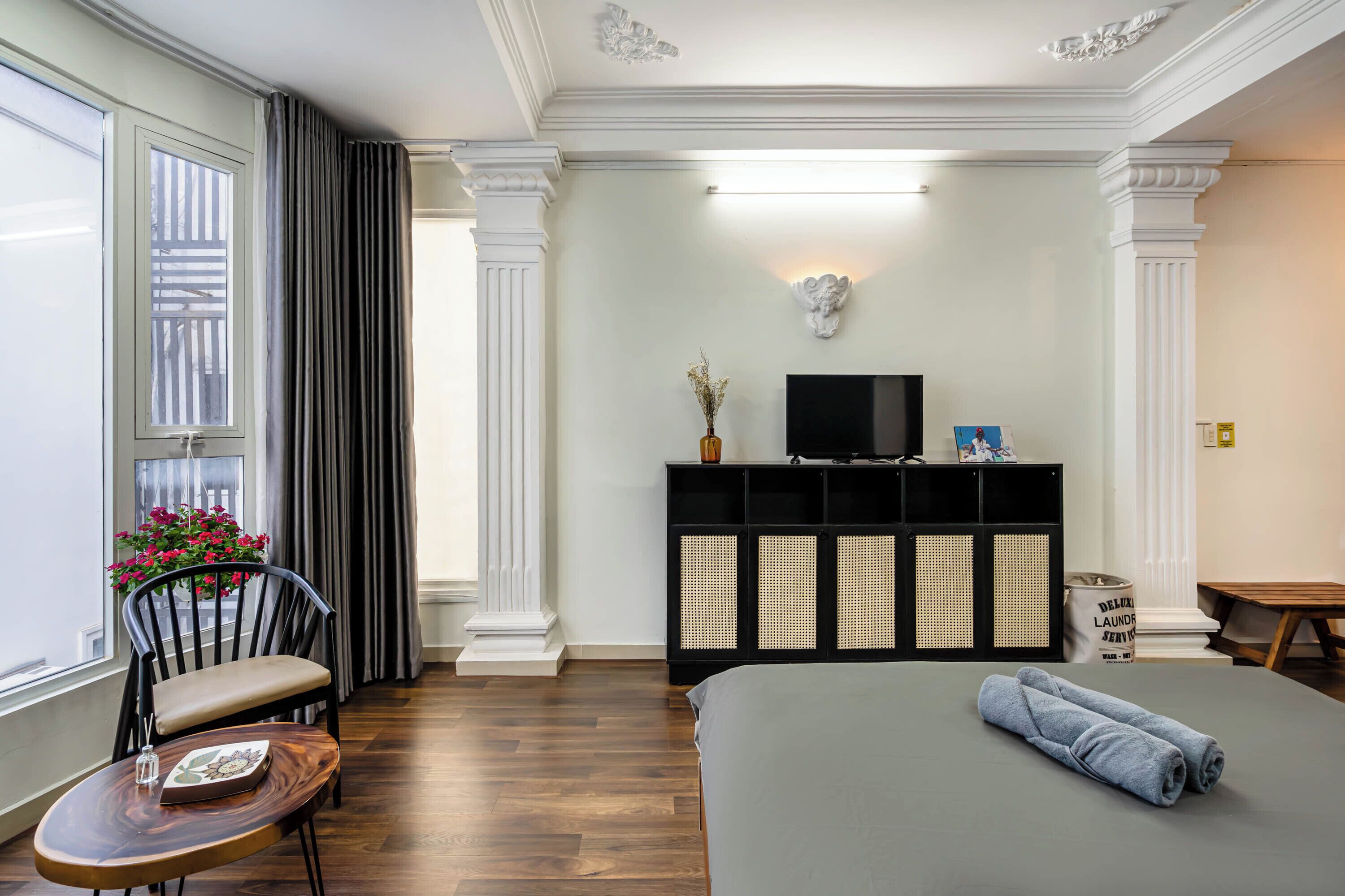 Habitación de hotel con cama, televisor, escritorio con silla, ventana con cortinas y estante montado en la pared con objetos decorativos.