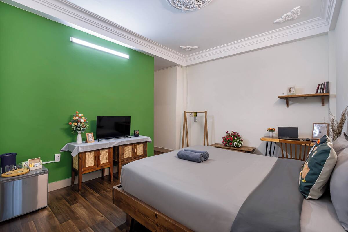 A hotel room with a green wall, a wooden bed frame, a flat-screen TV, a desk with a laptop, and a small shelf with books.