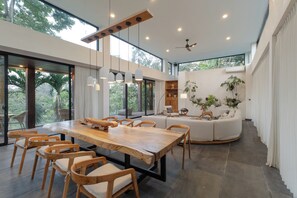 A modern dining area with a large wooden table and chairs, a ceiling fan, and large windows offering a view of the outdoors. - IBU The Sanctuary by OXO - Retreat House for Family & Transformational Events (Ubud)