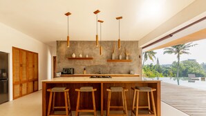 A modern kitchen with a central island, wooden stools, pendant lights, and a view of a tropical landscape. - IBU The Sanctuary by OXO - Retreat House for Family & Transformational Events (Ubud)