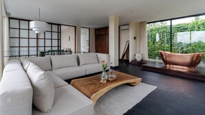 A modern living room with a sectional sofa, a wooden coffee table, and a large window with a view of greenery. - IBU The Sanctuary by OXO - Retreat House for Family & Transformational Events (Ubud)