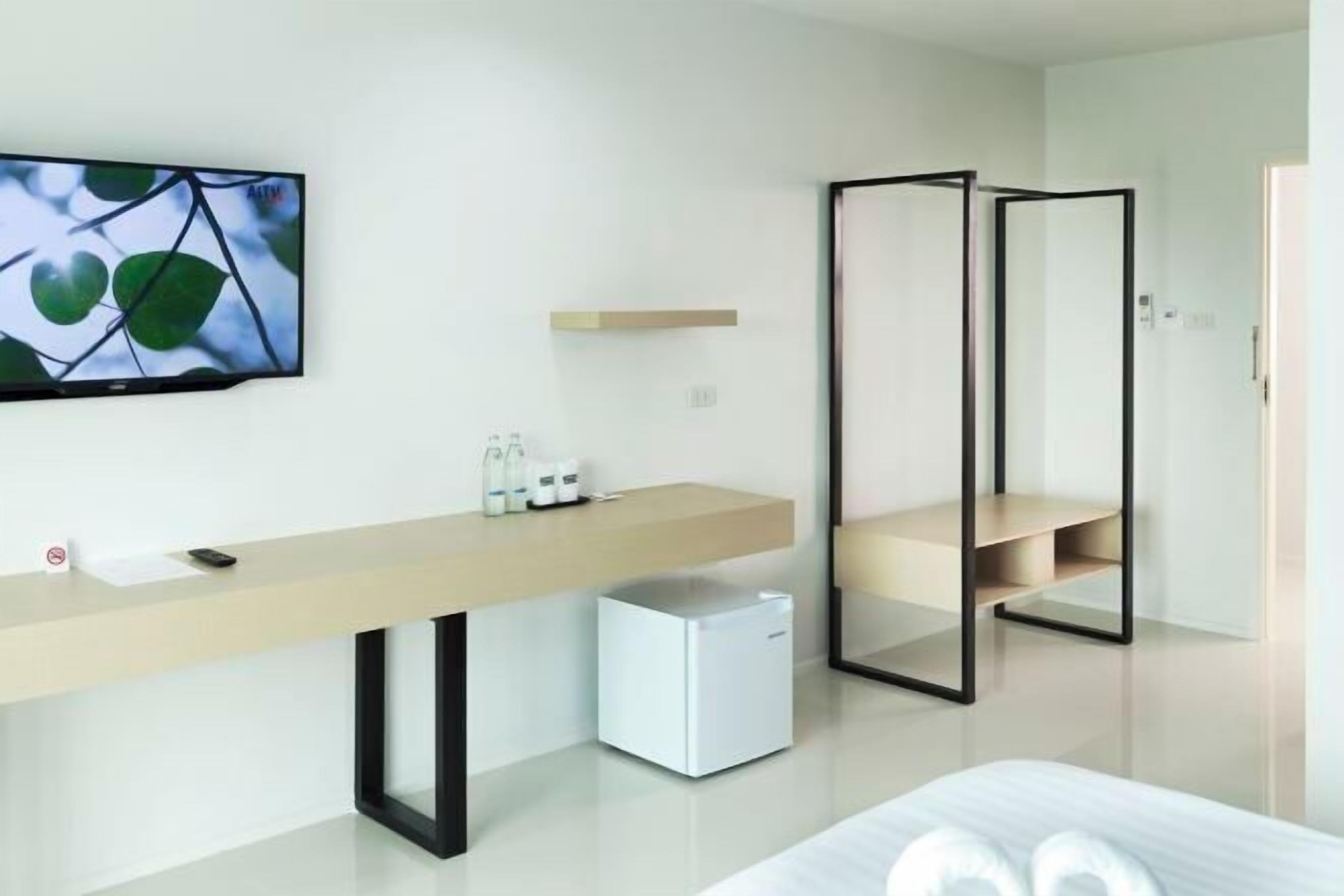 Una habitación de hotel moderna con diseño minimalista, que incluye un escritorio de madera alargado, un refrigerador blanco pequeño, una cama con ropa blanca y un espejo grande.
