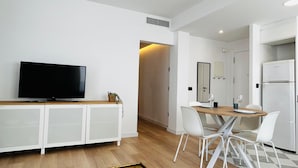 Een moderne, minimalistische woonkamer met een flatscreen-tv, een houten eettafel, witte stoelen en een witte kast.