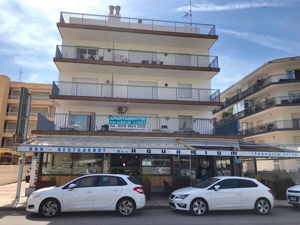 En bygning med flere etager, altaner, en restaurant der hedder "BAR RESTAURANTE", og et skilt, der annoncerer lejligheder til leje.