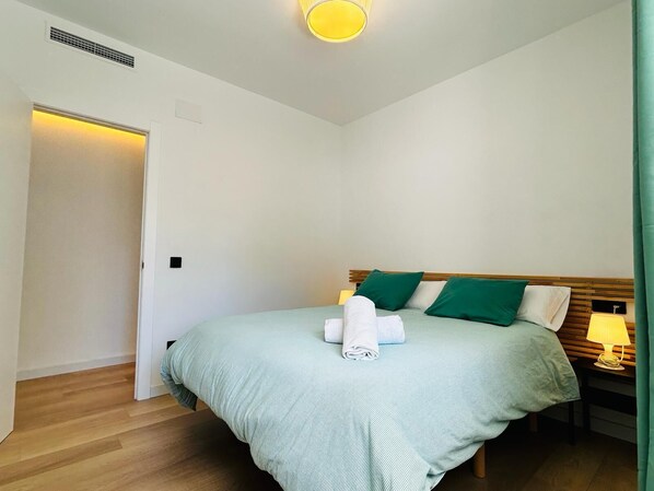 A neatly made bed with a green bedspread and pillows, a bedside lamp, and a rolled white towel on the bed. - APARTAMENTOS AQUARIUM. (Roses)