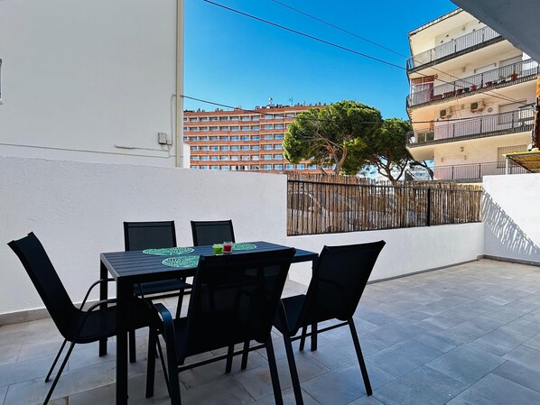 A terrace with a table and chairs, a view of a building, and a tree. - APARTAMENTOS AQUARIUM. (Roses)