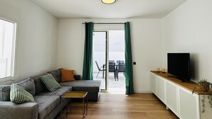 Een moderne woonkamer met een grijze bank, een klein bijzettafeltje, een televisie op een kast en uitzicht op buiten.