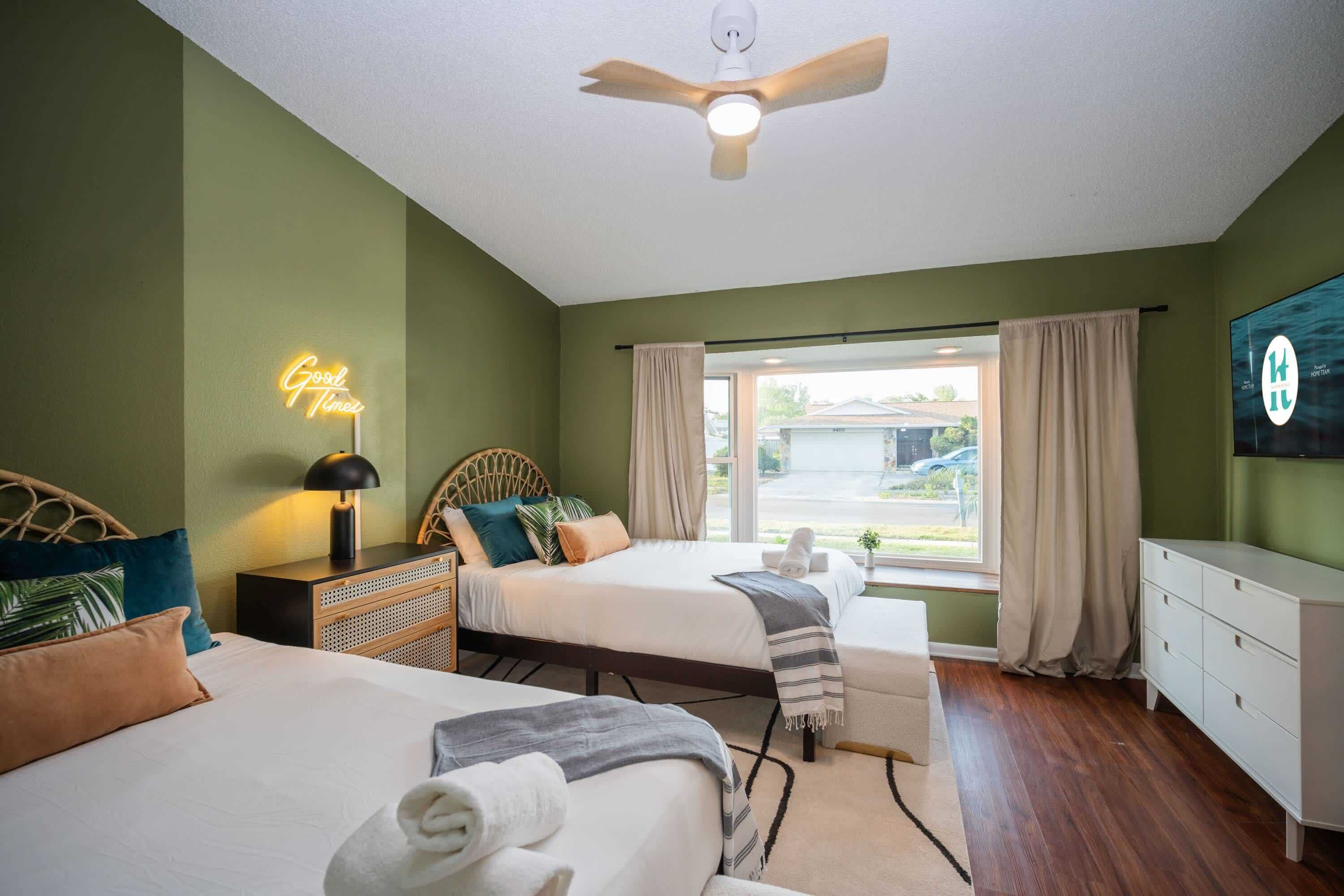 A hotel room with two beds, a ceiling fan, a neon sign, and a window with curtains.