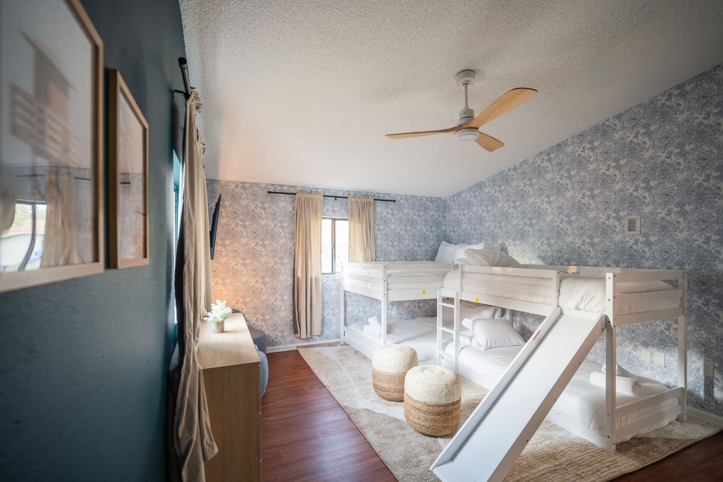 A bedroom with a bunk bed, a ceiling fan, a window with curtains, and a wooden floor.