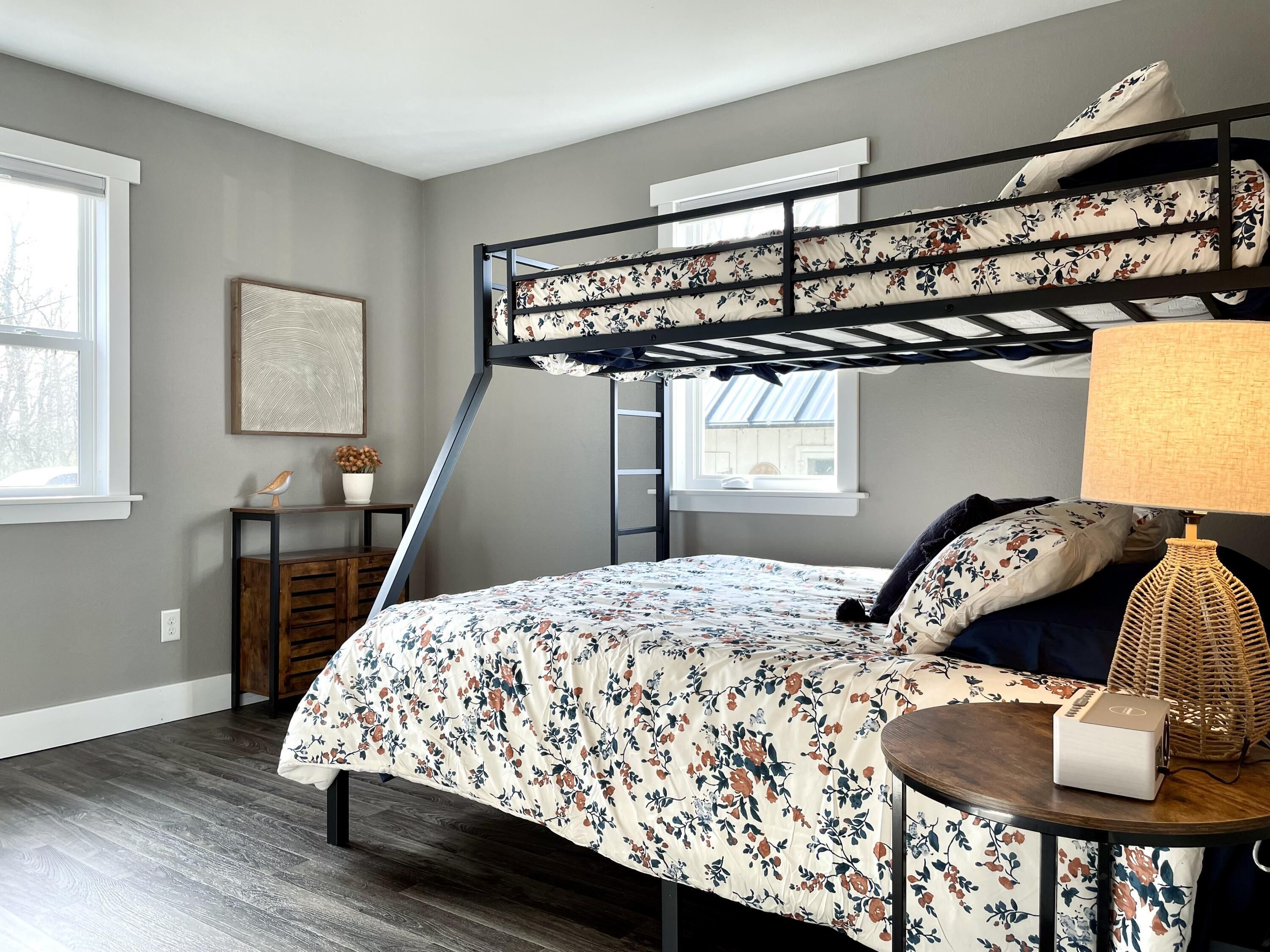A bedroom with a bunk bed, a bedside table with a lamp, a small desk with a plant, and a framed picture on the wall.