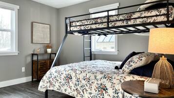 A bedroom with a bunk bed, a bedside table with a lamp, a small desk with a plant, and a framed picture on the wall.